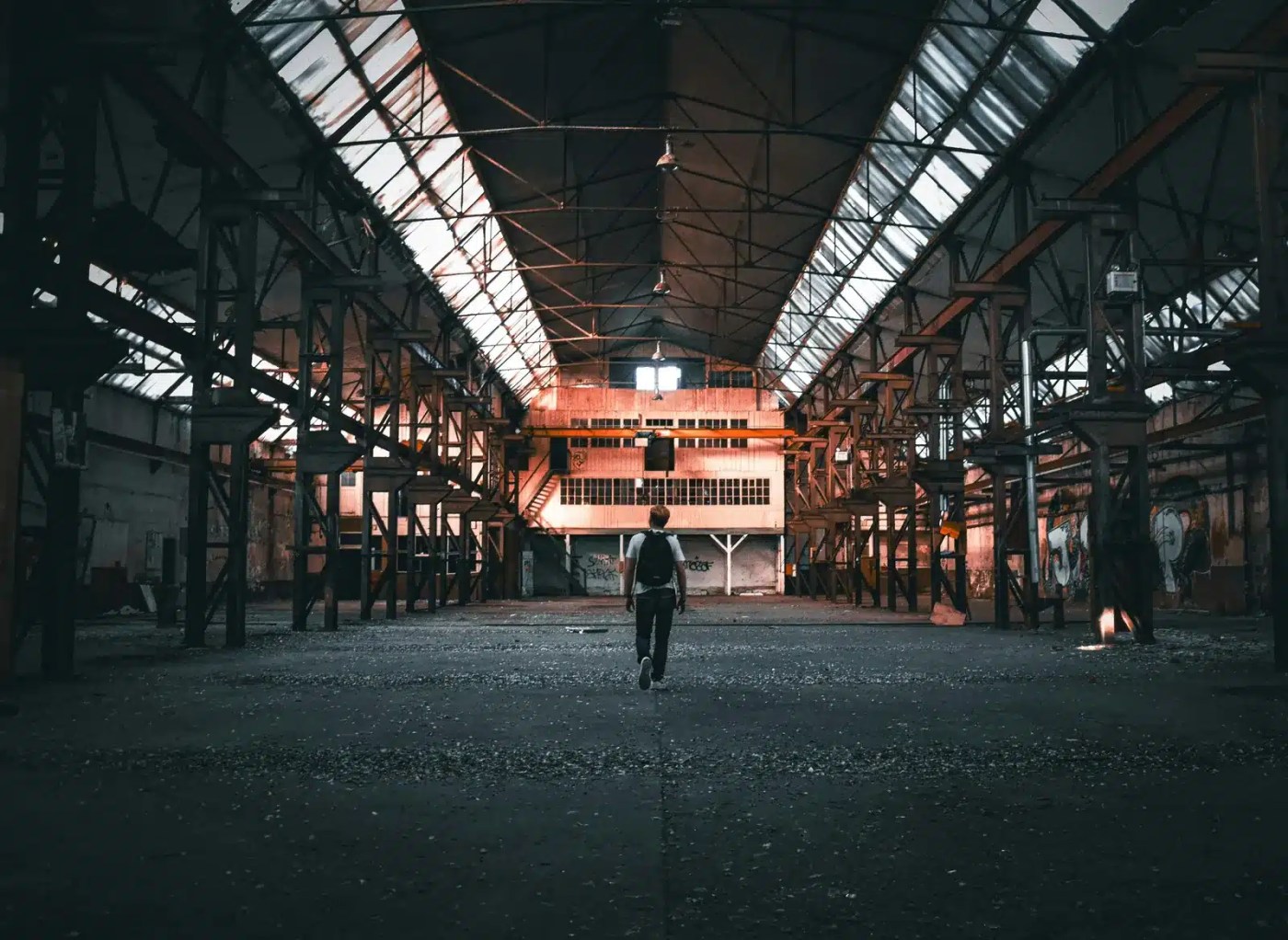 Worker with a backpack walking alone through a vast, empty industrial hall at the end of his shift
